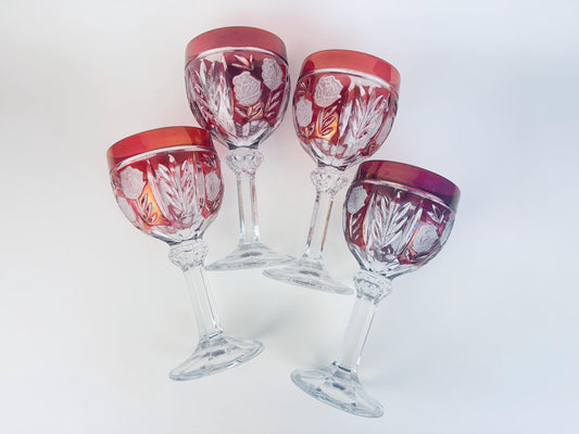 Set of four vintage ruby-flash cut glass wine goblets with frosted rose and leaf panels, displayed angled on a white background to show the deep red rims and clear pressed stems.