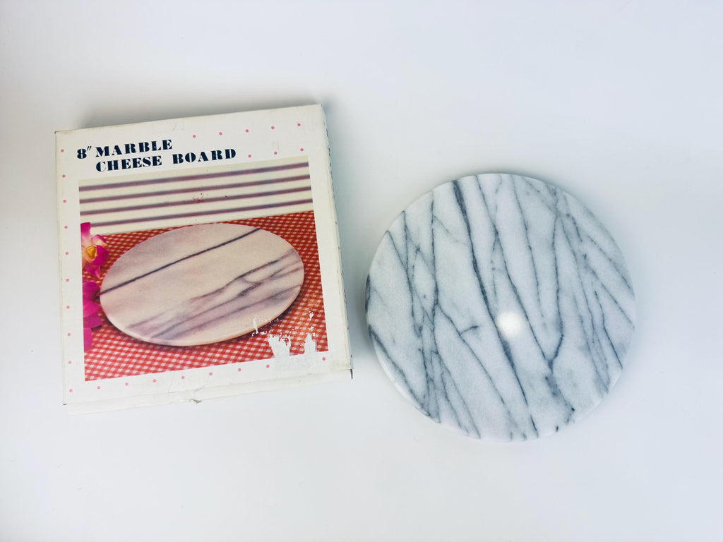 Vintage 8-inch round white and grey veined marble cheese board shown beside its original 1980s box, which features a printed product photo and pink polka-dot border. The marble board has a smooth polished surface with linear grey striations.