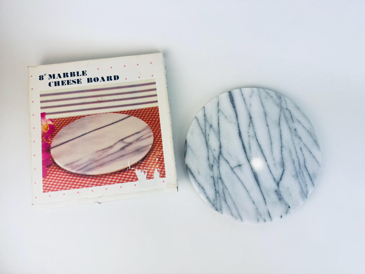 Vintage 8-inch round white and grey veined marble cheese board shown beside its original 1980s box, which features a printed product photo and pink polka-dot border. The marble board has a smooth polished surface with linear grey striations.