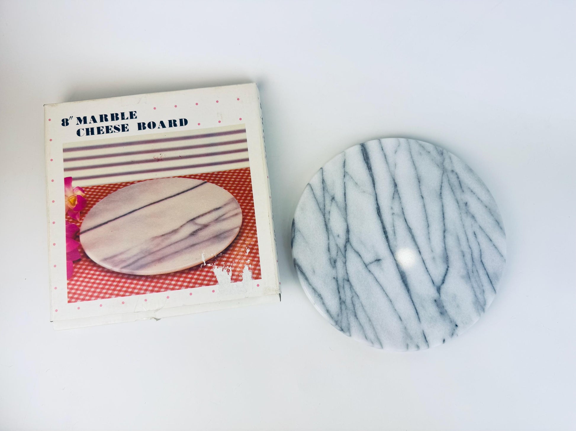 Vintage 8-inch round white and grey veined marble cheese board shown beside its original 1980s box, which features a printed product photo and pink polka-dot border. The marble board has a smooth polished surface with linear grey striations.