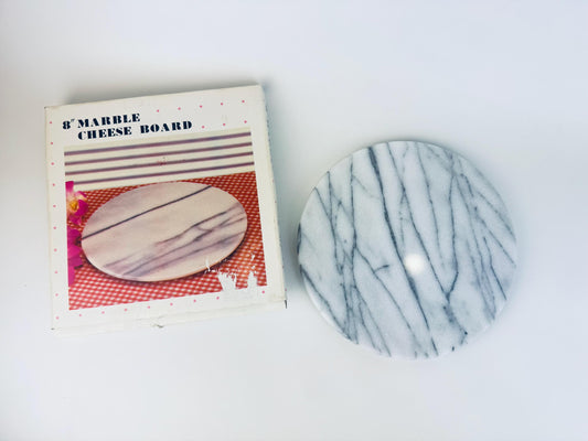 Vintage 8-inch round white and grey veined marble cheese board shown beside its original 1980s box, which features a printed product photo and pink polka-dot border. The marble board has a smooth polished surface with linear grey striations.