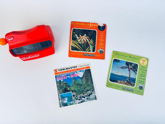 Red vintage GAF View-Master viewer displayed alongside three Hawaii-themed View-Master reel packets—‘Flowers of Hawaii,’ ‘Hawaiian Medley: Life in Aloha Land,’ and ‘Kauai and Maui’ from the United States Travel series—arranged on a clean white background.
