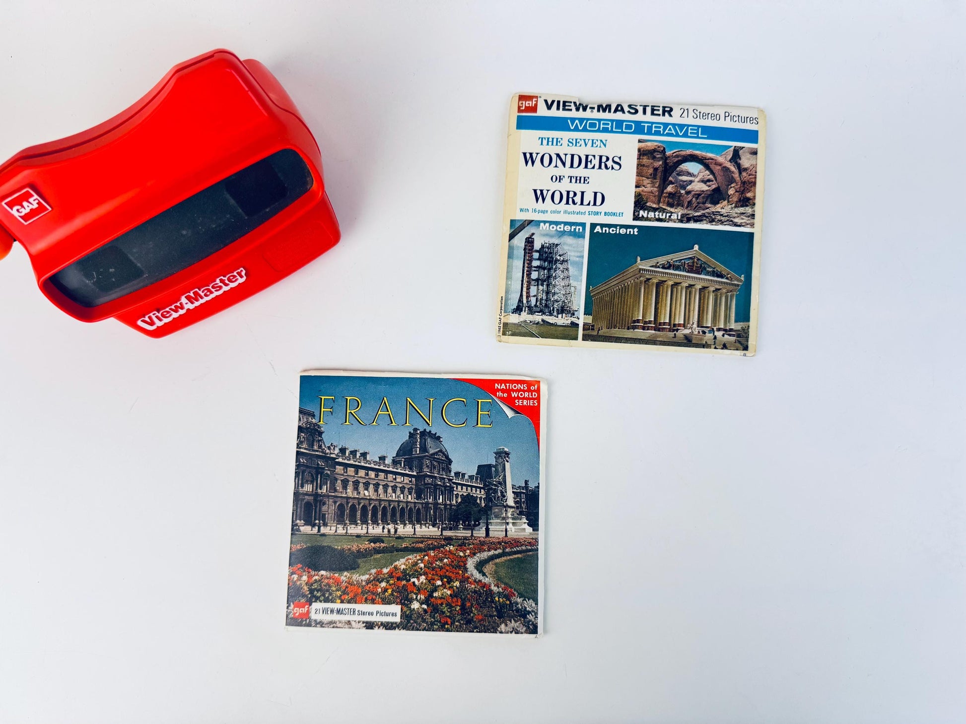 Red vintage View-Master viewer displayed with two 1960s–1970s GAF View-Master reel packets—‘Seven Wonders of the World’ and ‘France’ from the Nations of the World series—arranged on a clean white background.