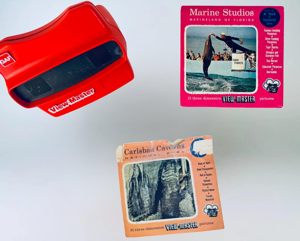 Red vintage View-Master viewer photographed alongside two 1960s–1970s View-Master reel packets, including Marine Studios Marineland of Florida and Carlsbad Caverns National Park, displayed against a soft grey background.