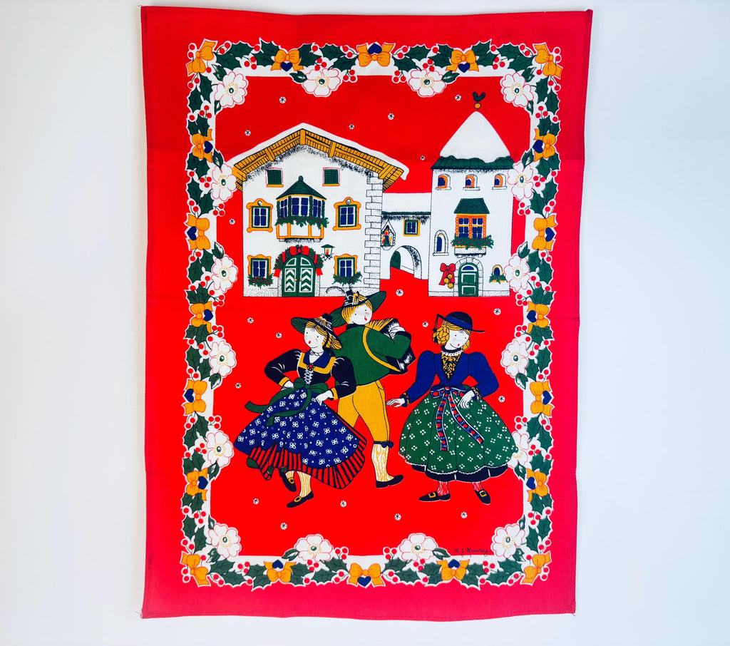 Vintage 1980s Italian cotton kitchen towel by M.J. Maurey featuring a bright red background with an Alpine village scene and three folk-dressed dancers framed by holly, berries and floral borders.