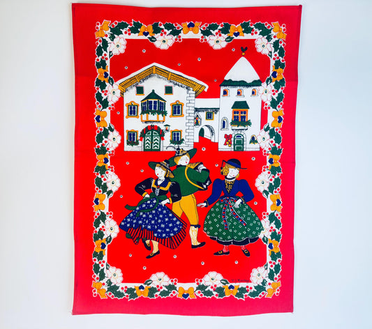 Vintage 1980s Italian cotton kitchen towel by M.J. Maurey featuring a bright red background with an Alpine village scene and three folk-dressed dancers framed by holly, berries and floral borders.
