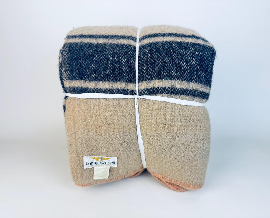Folded vintage beige wool-blend camp blanket with indigo stripes, secured with white ties, featuring a mid-century Hercules label on the corner.