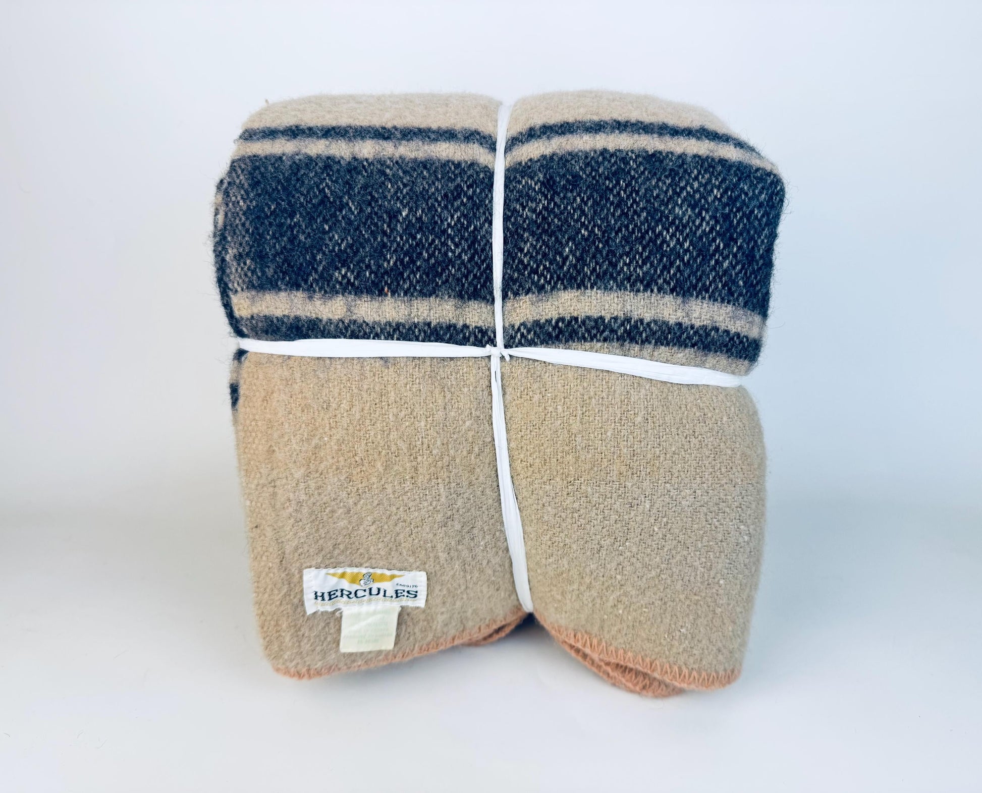 Folded vintage beige wool-blend camp blanket with indigo stripes, secured with white ties, featuring a mid-century Hercules label on the corner.