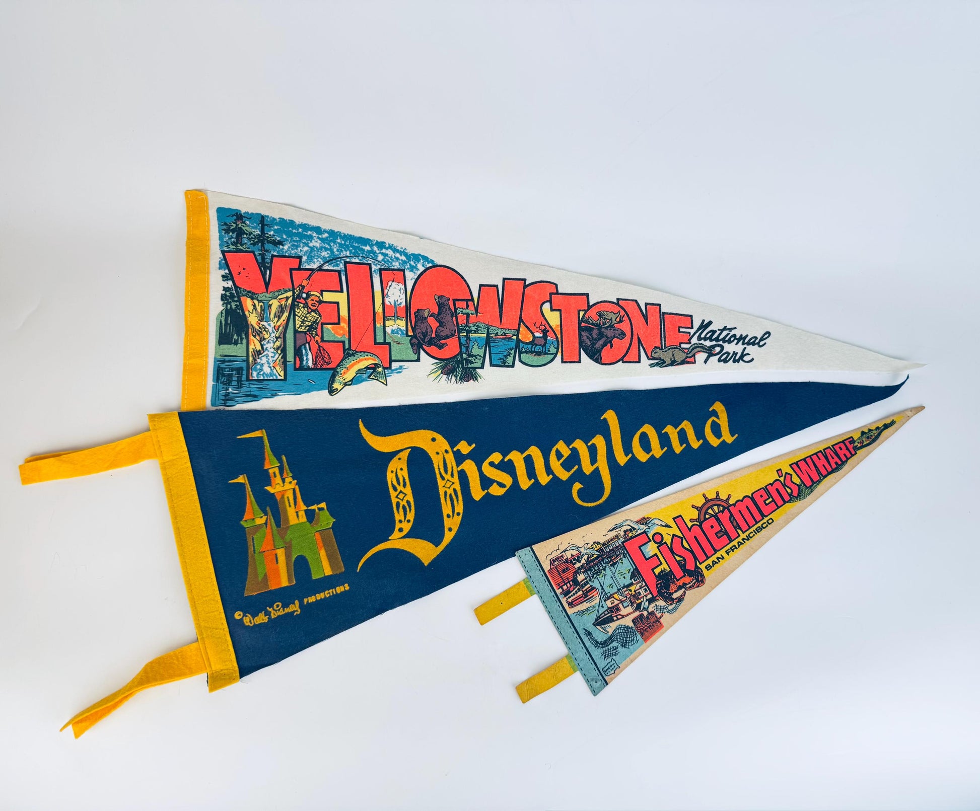 Three vintage mid-century felt souvenir pennants—Yellowstone National Park, Disneyland, and Fisherman’s Wharf—layered on a white background, each featuring bold retro graphics and colourful printed designs.