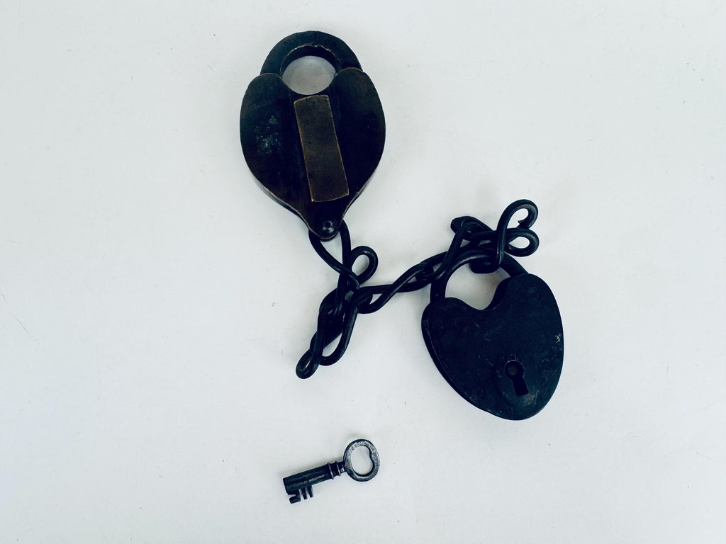 Close-up of a heavy, heart-shaped 1870s brass and iron railway padlock with original key inserted, showing patinated metal and worn keyhole, set on white background.