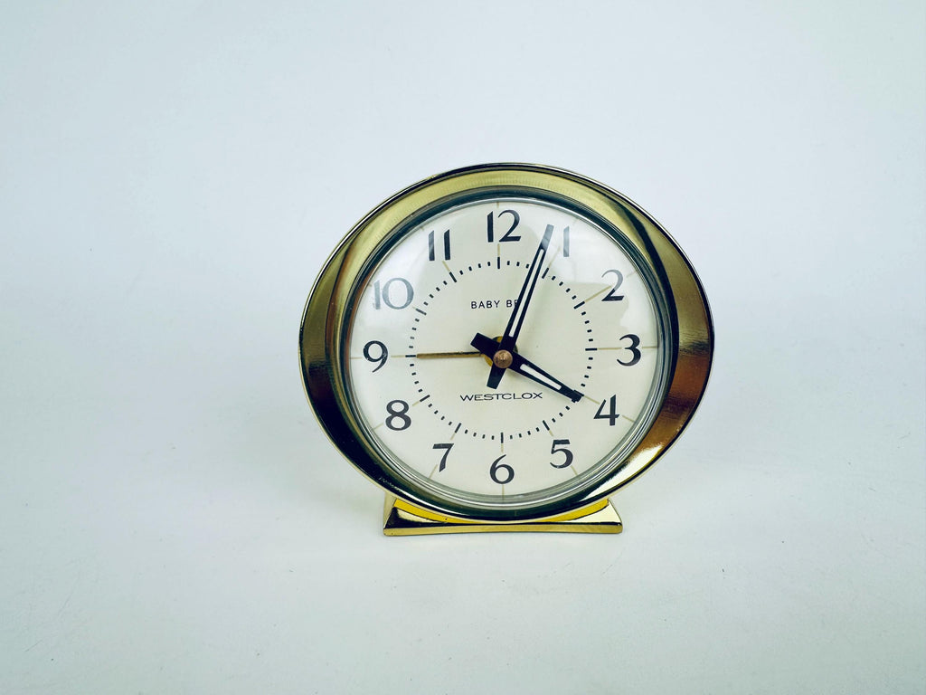 Gold-tone brass style Baby Ben alarm clock on white background — mid-size round face with black numerals and matching gold-tone base.