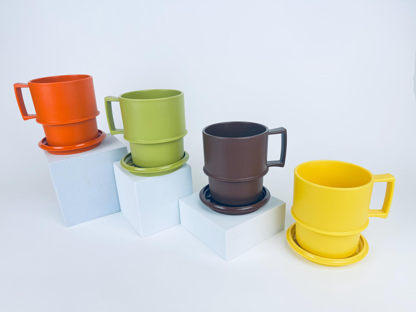 Four vintage 1970s Tupperware stackable mugs in orange, green, brown, and yellow, each shown with matching coaster-lids, arranged on white pedestals.