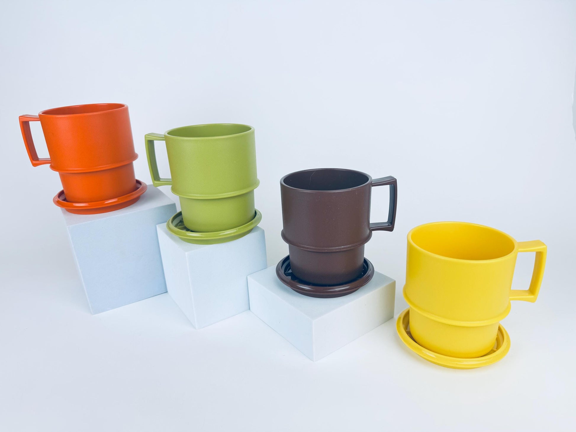 Four vintage 1970s Tupperware stackable mugs in orange, green, brown, and yellow, each shown with matching coaster-lids, arranged on white pedestals.