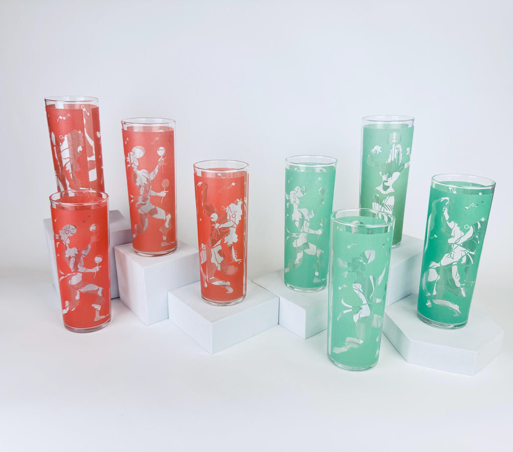 Eight vintage mid-century tall highball glasses — four coral and four mint — each decorated with a white frosted stylized flamenco dancer motif, displayed on white minimalist pedestals against a white background.