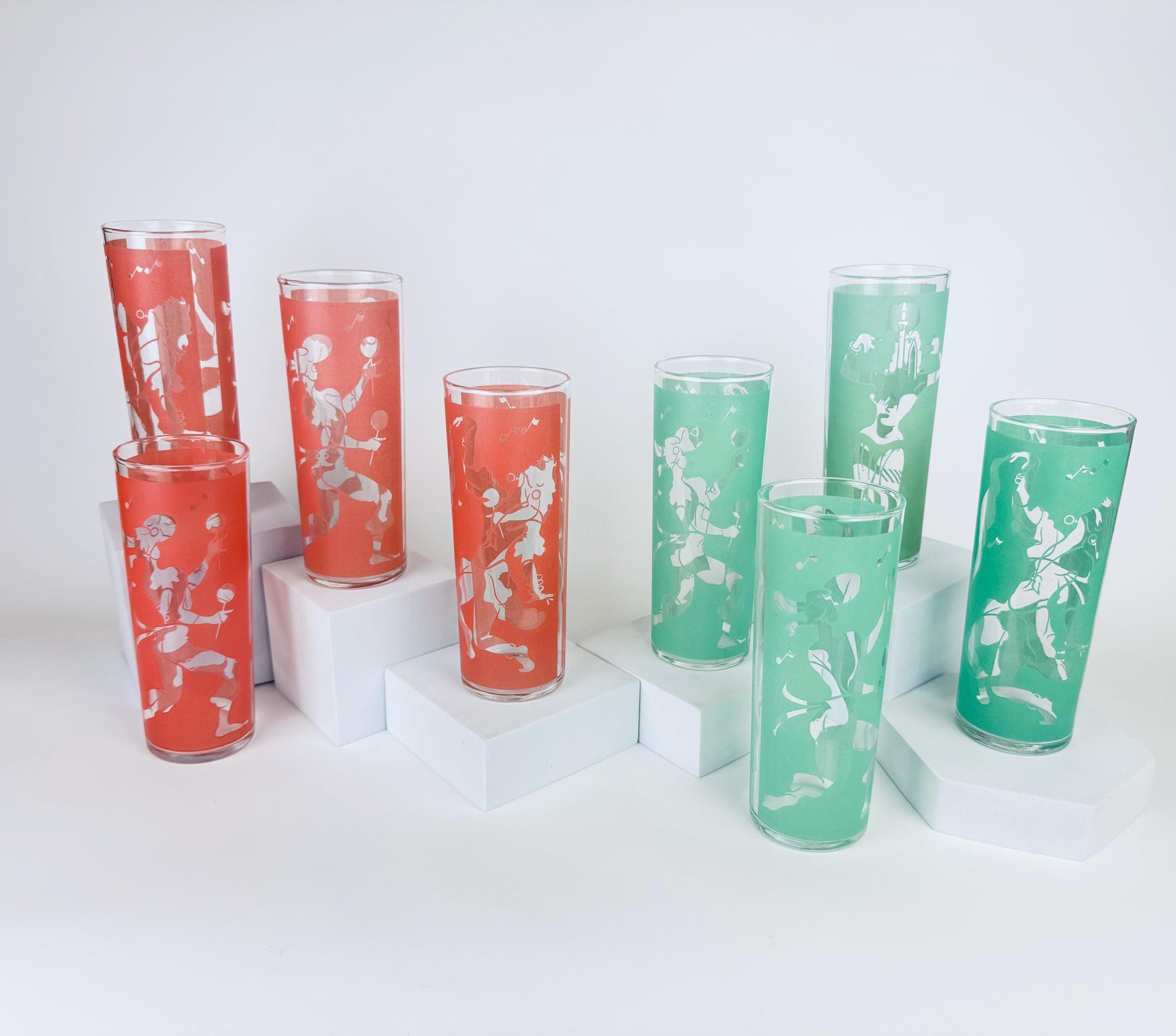 Eight vintage mid-century tall highball glasses — four coral and four mint — each decorated with a white frosted stylized flamenco dancer motif, displayed on white minimalist pedestals against a white background.