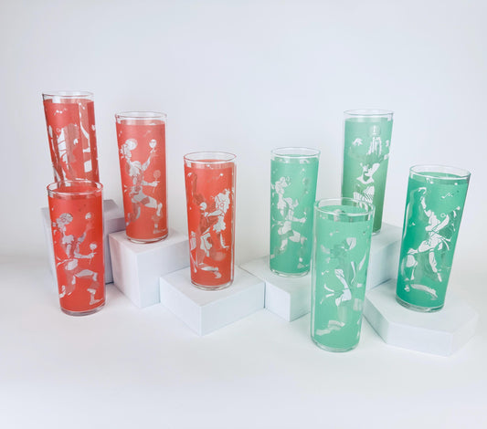Eight vintage mid-century tall highball glasses — four coral and four mint — each decorated with a white frosted stylized flamenco dancer motif, displayed on white minimalist pedestals against a white background.